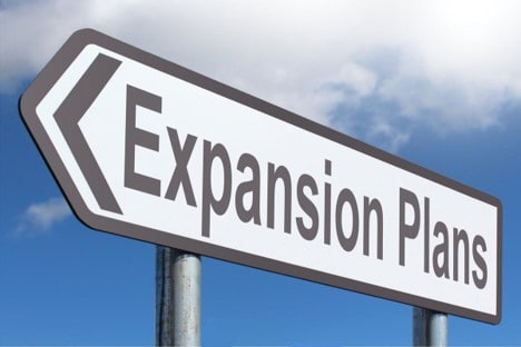  A white and grey sign that says Expansion Plans set against a sky blue and cloud background.